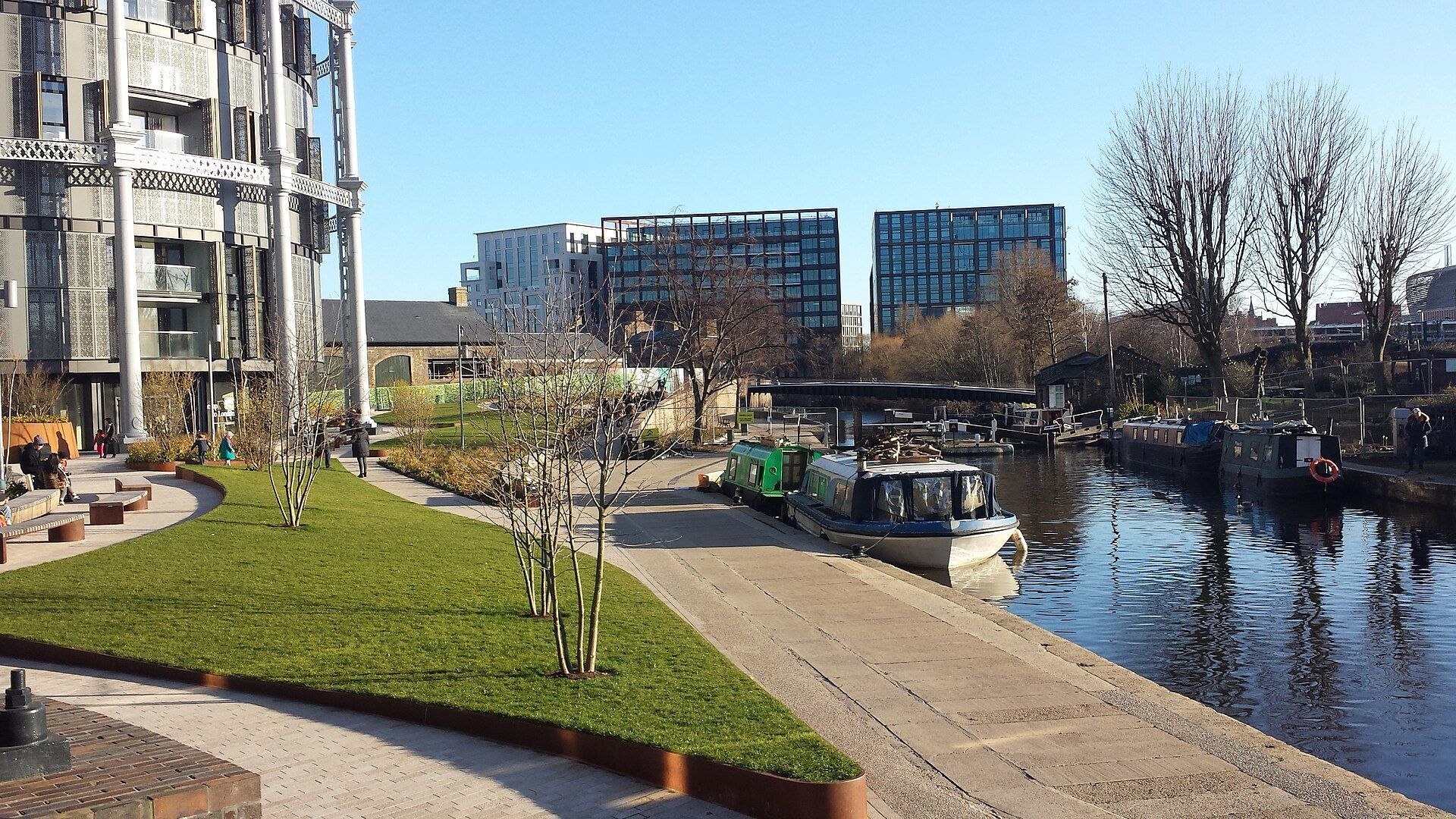 A canal with boats and modern buildings on a sunny day. Green grass and trees are visible.