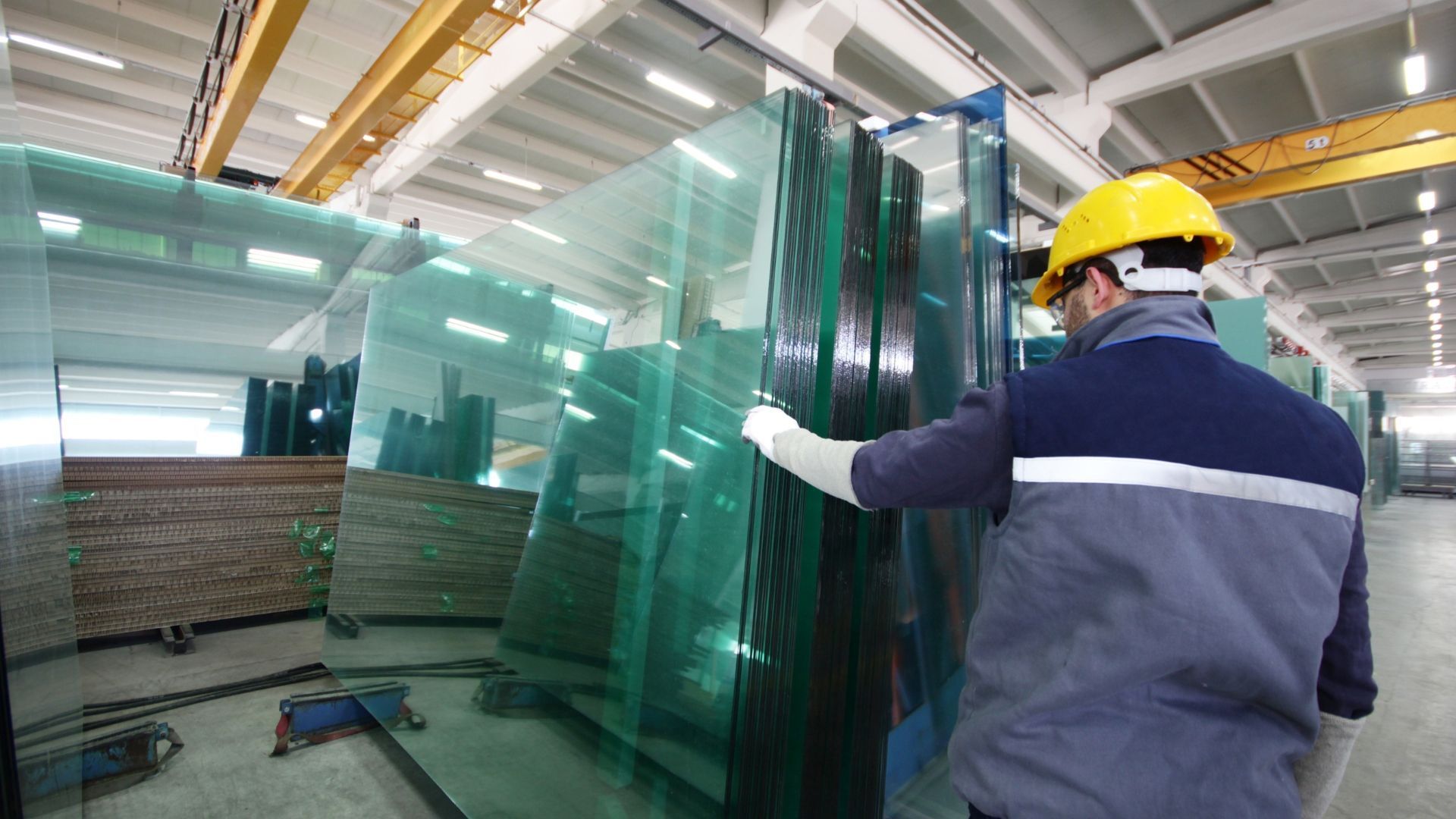 Worker wearing a hard hat and gloves handling large glass sheets in a factory.