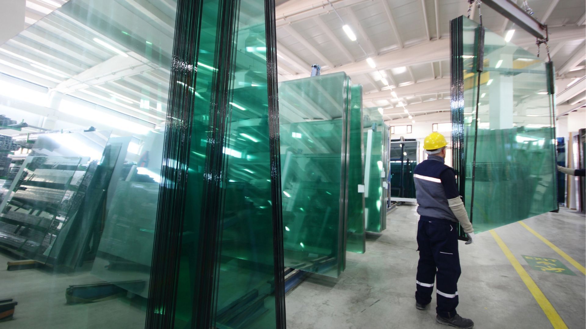 Worker in a factory inspecting large sheets of green glass, possibly for construction.