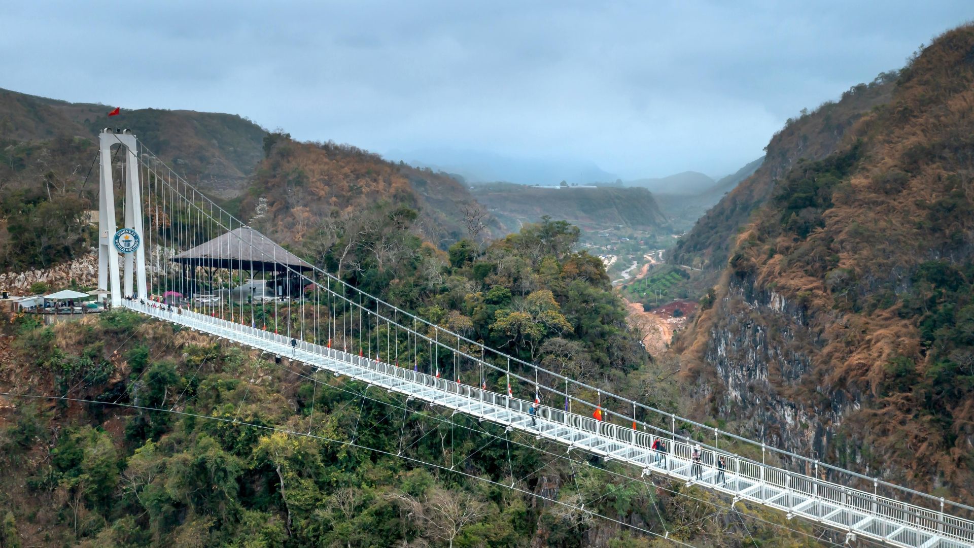A long glass-bottomed bridge spans a valley, with people walking on it. Mountains surround the bridge.