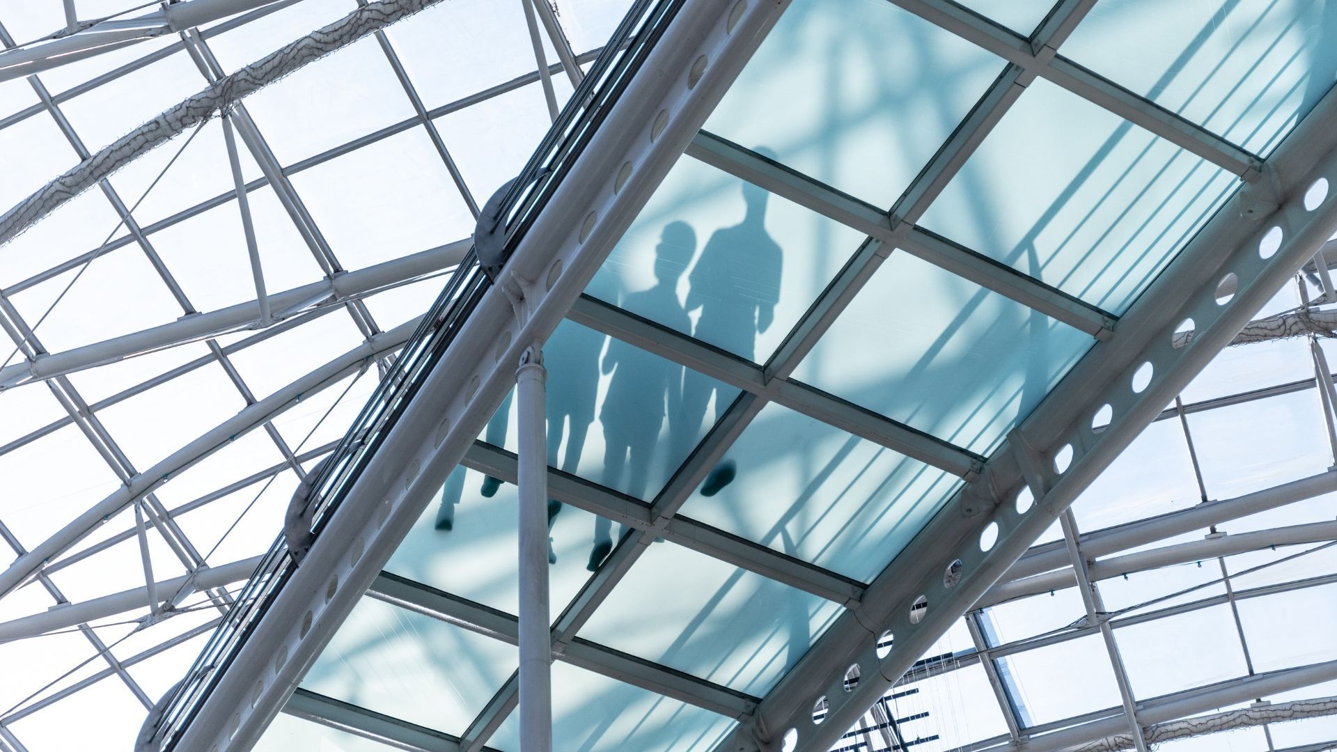 Silhouettes of three people walking on a glass-bottomed walkway under a metal grid roof.