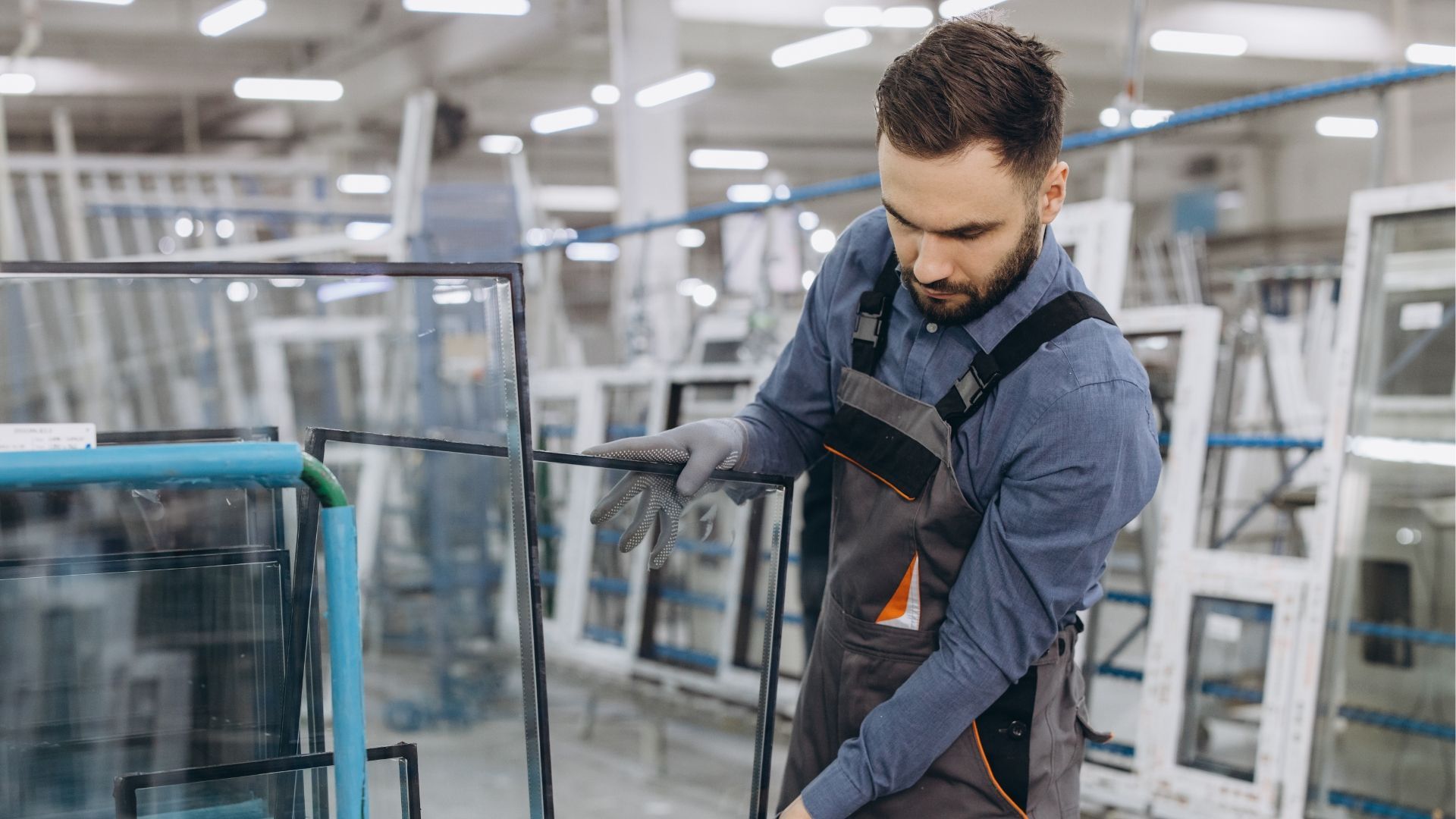 Man working with glass in a window manufacturing factory, wearing gloves and overalls.