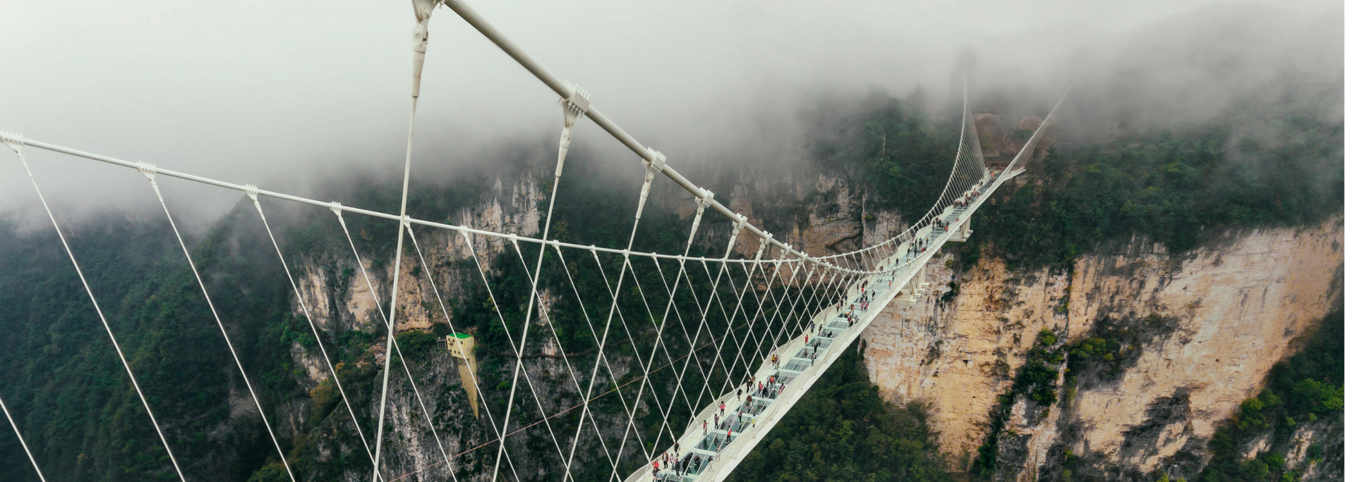A long glass-bottomed suspension bridge with steel cables over a foggy canyon.