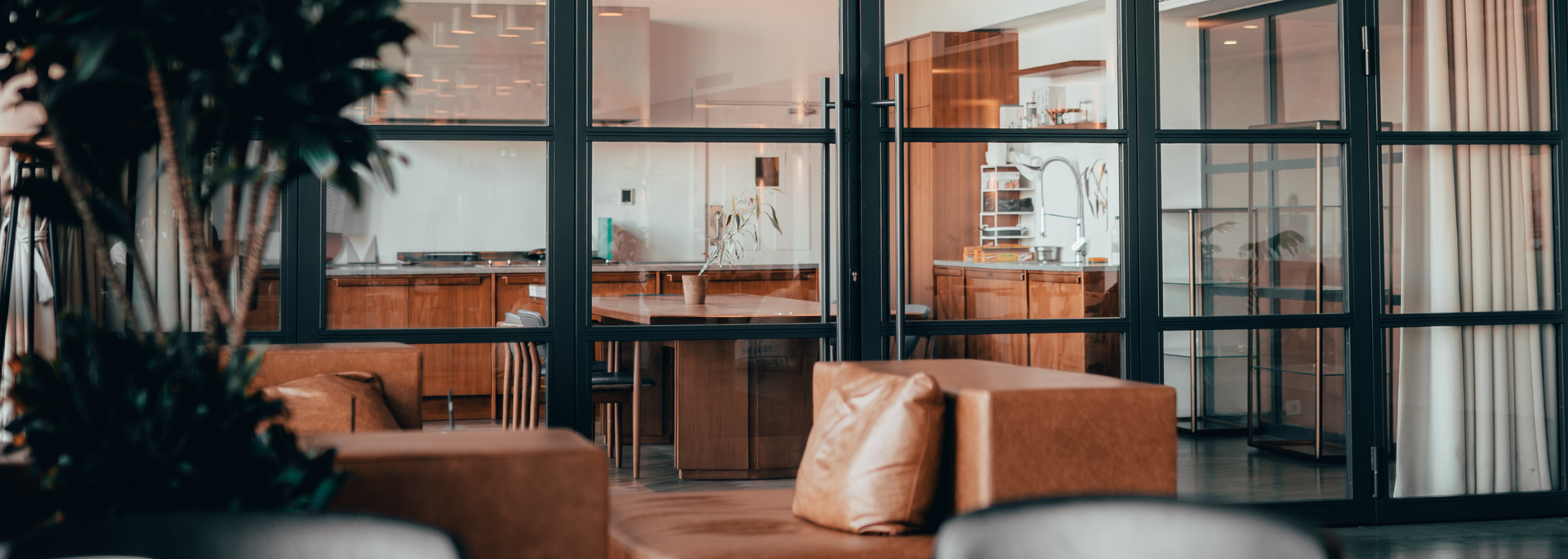 Interior view of a modern space with glass doors. The kitchen is visible through the doors. Brown sofas are in the foreground.