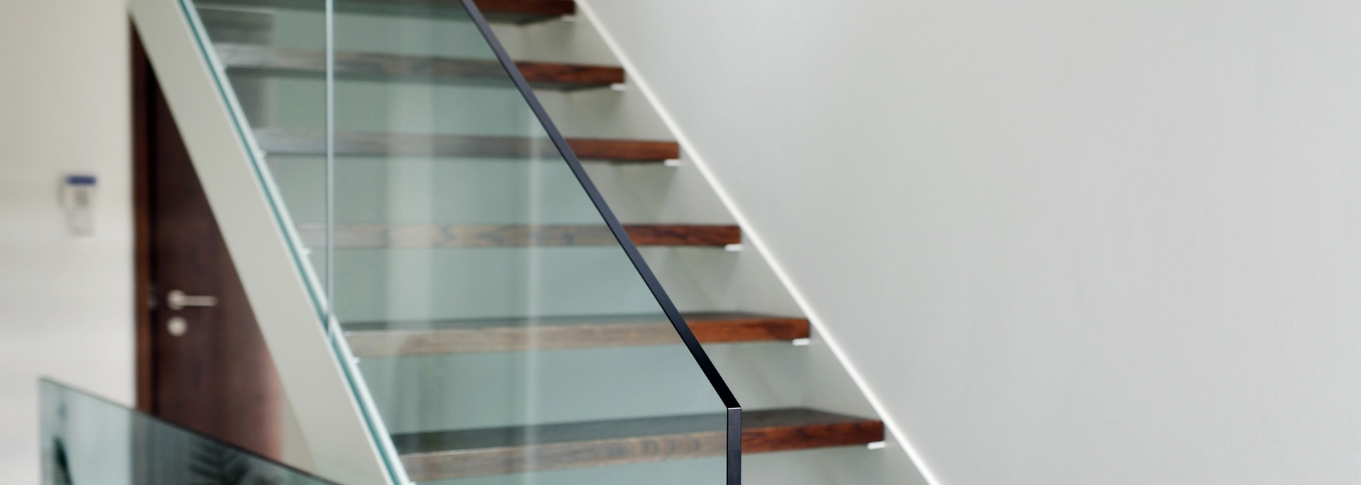 Staircase with wooden steps and glass railing. A black handrail runs along the stairs against a white wall.