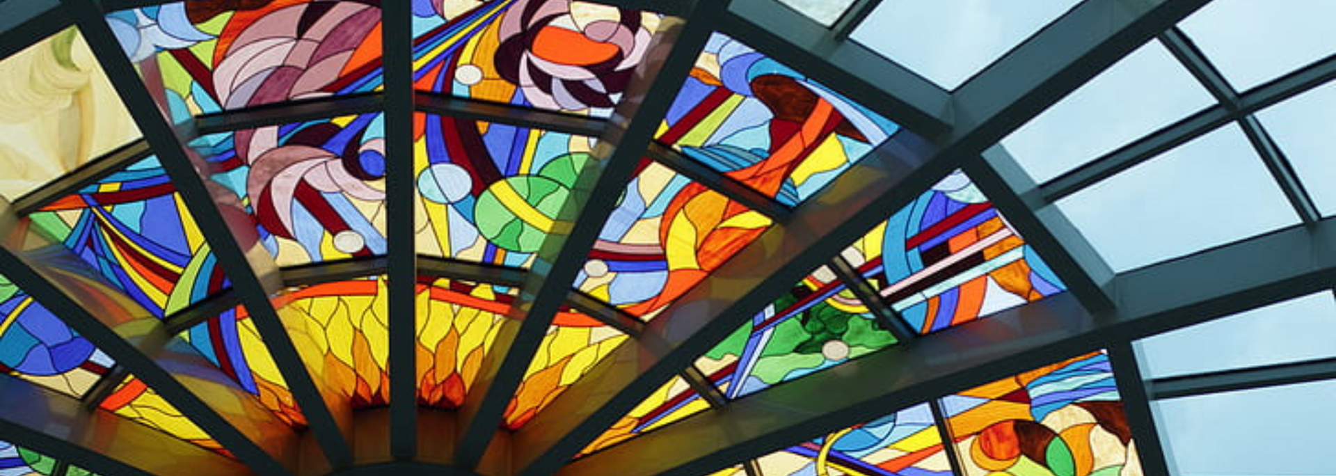 Stained-glass window with colorful abstract design, framed by dark metal. Light blue sky visible.