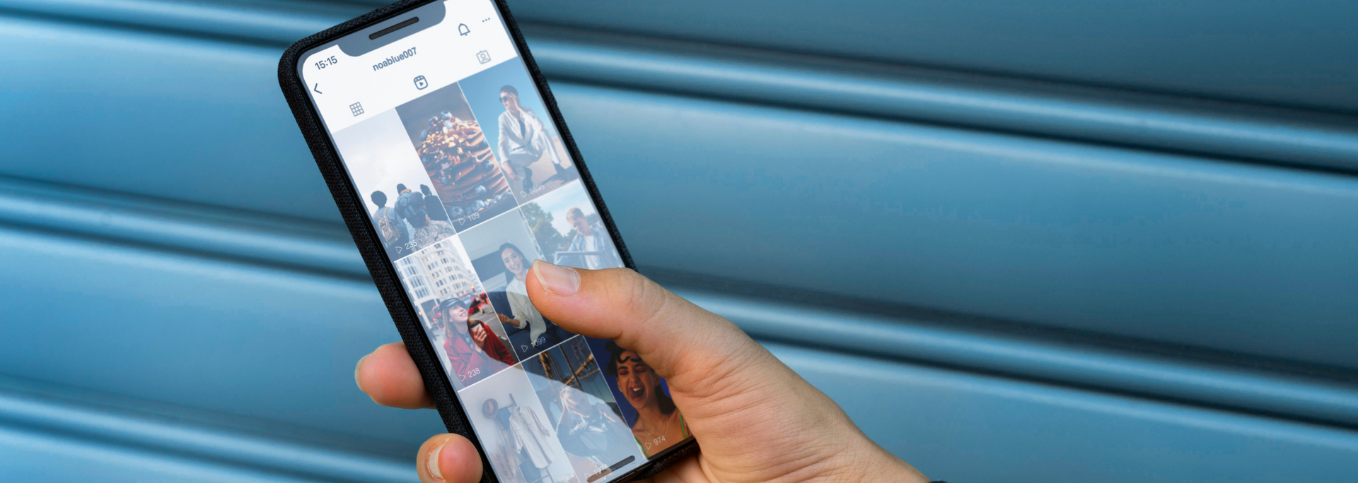 A person's hand holding a smartphone, scrolling on a social media app, set against a blue wall.