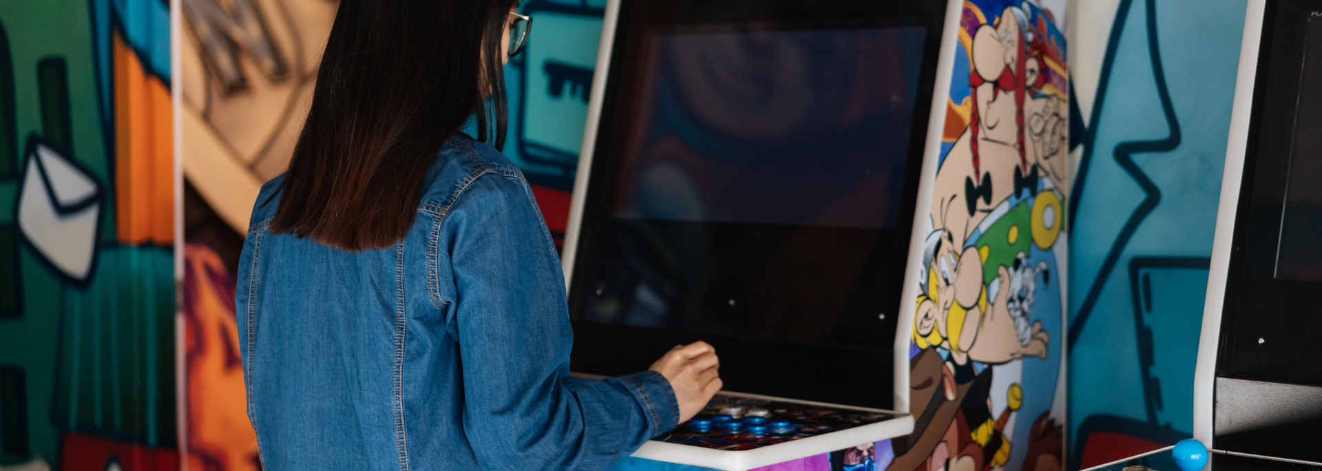 Person playing a video game at an arcade, with a jean jacket and colorful wall art.