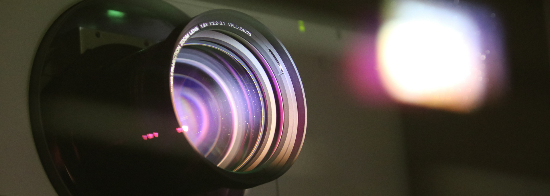 Close-up of a projector lens with light projecting onto a surface; lens has purple and yellow colors.