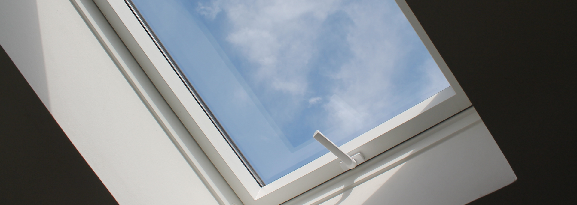 A white skylight angled towards a blue sky with white clouds. A white handle is visible.