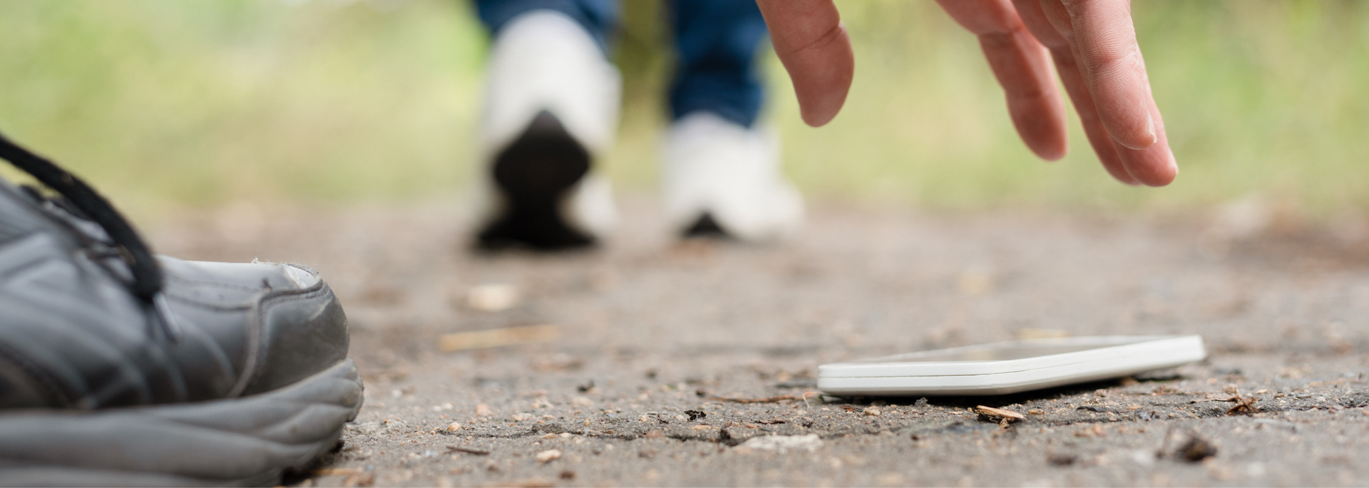 A hand reaching for a dropped white cell phone on a walkway with feet in the background.