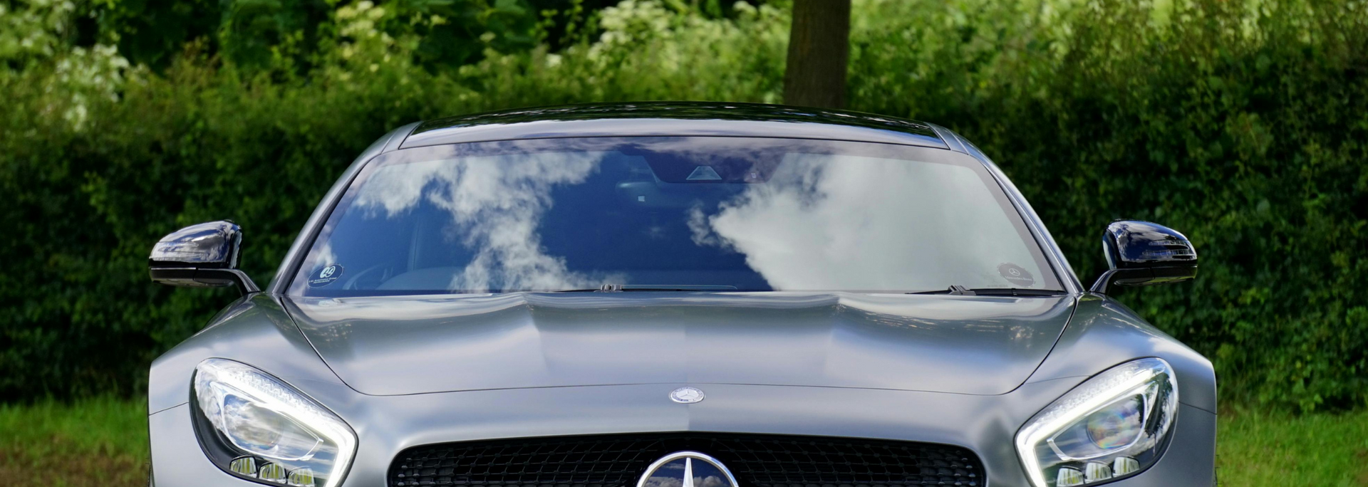A gray Mercedes-Benz car, front view, with the windshield reflecting the sky, parked in front of green foliage.