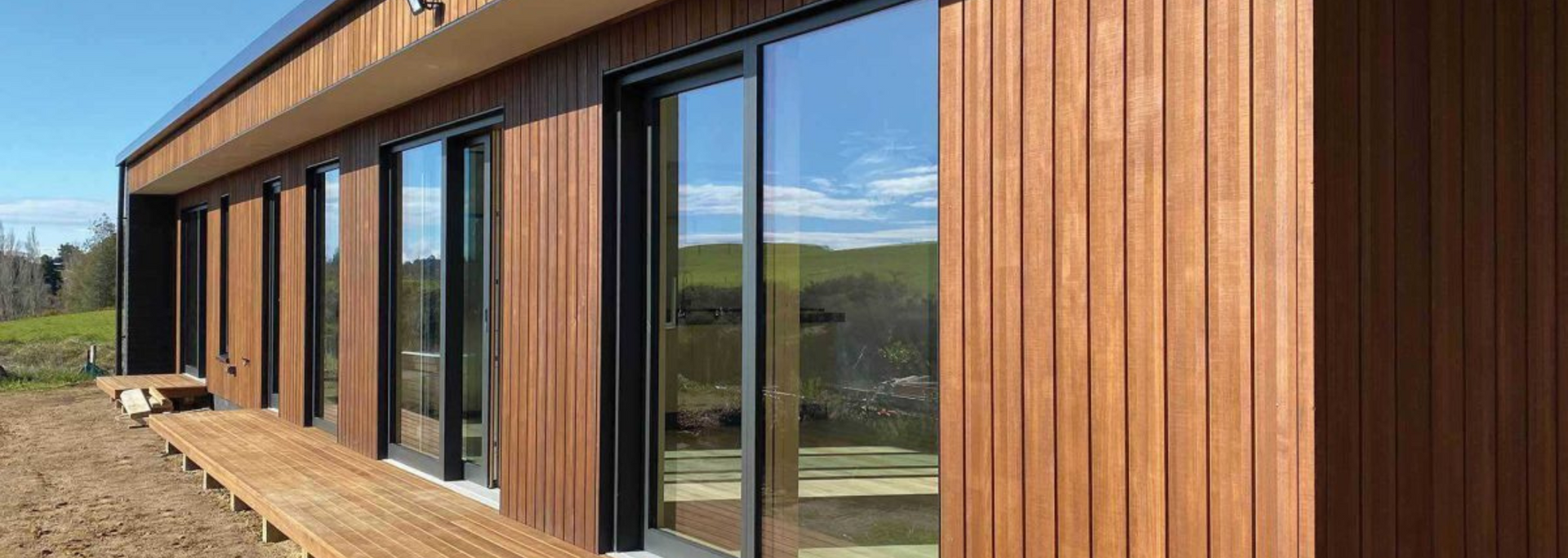 Exterior of a modern building with wooden siding and large glass windows reflecting a blue sky and green hills.