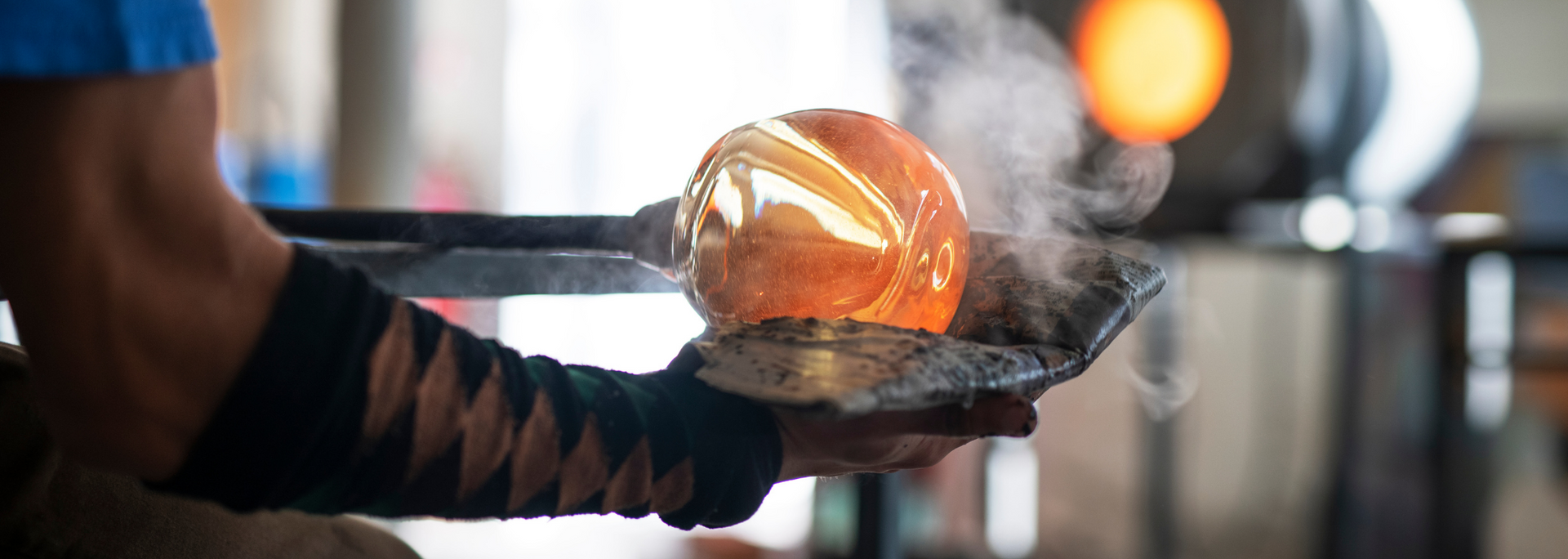 Glassblower holding a glowing, molten glass sphere with steam rising.