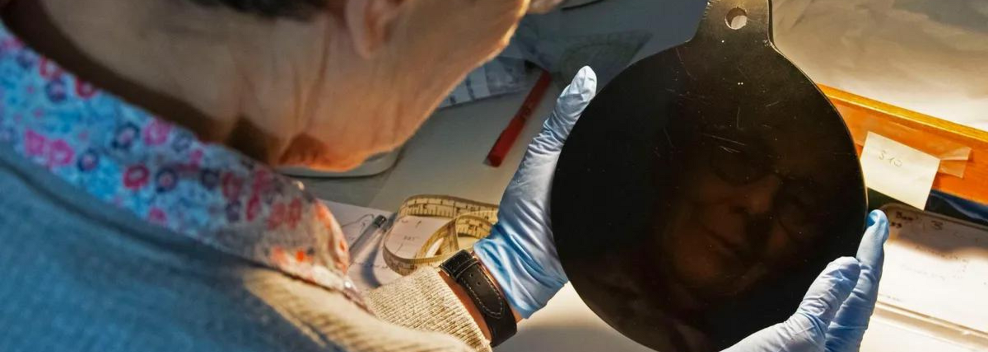 Person wearing blue gloves holding a dark, round object; a face is reflected in the object.