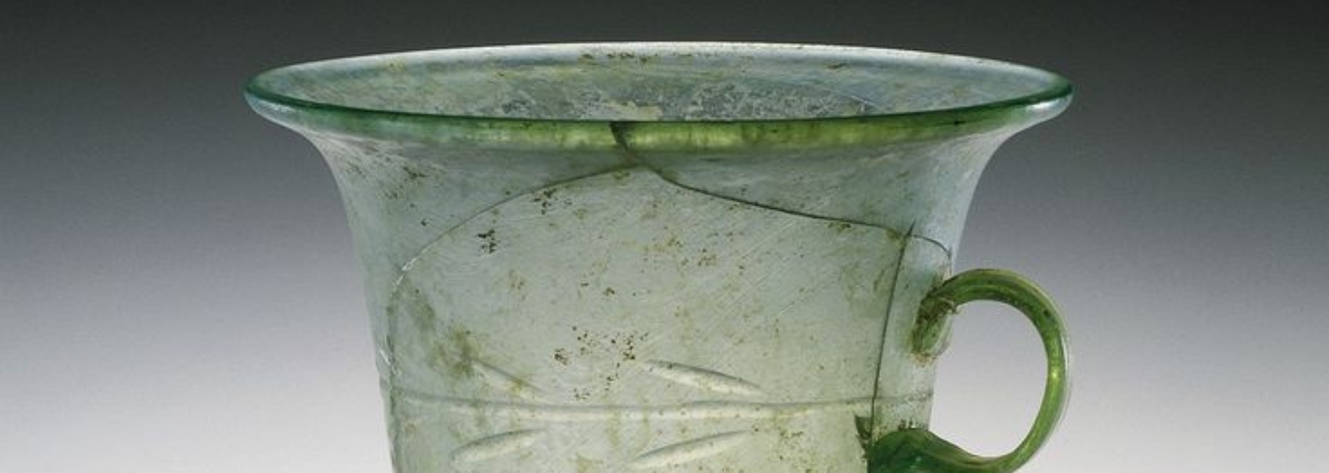 A pale green glass mug with a handle and decorative etched lines. It has cracks and is on a gray background.