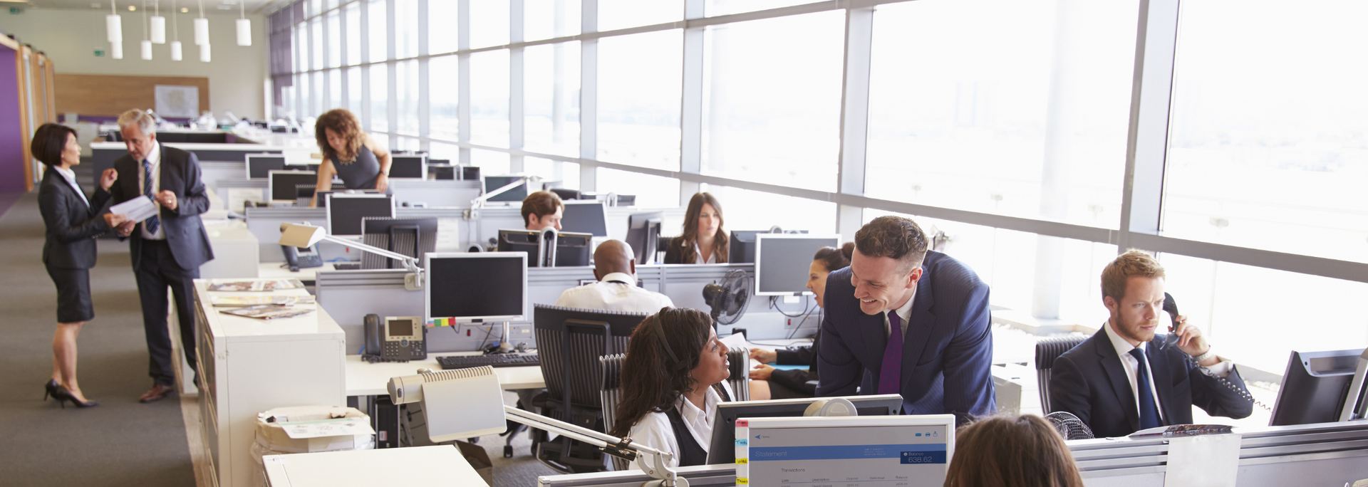 People working in an open-plan office setting; some in suits; some on the phone; large windows in the background.