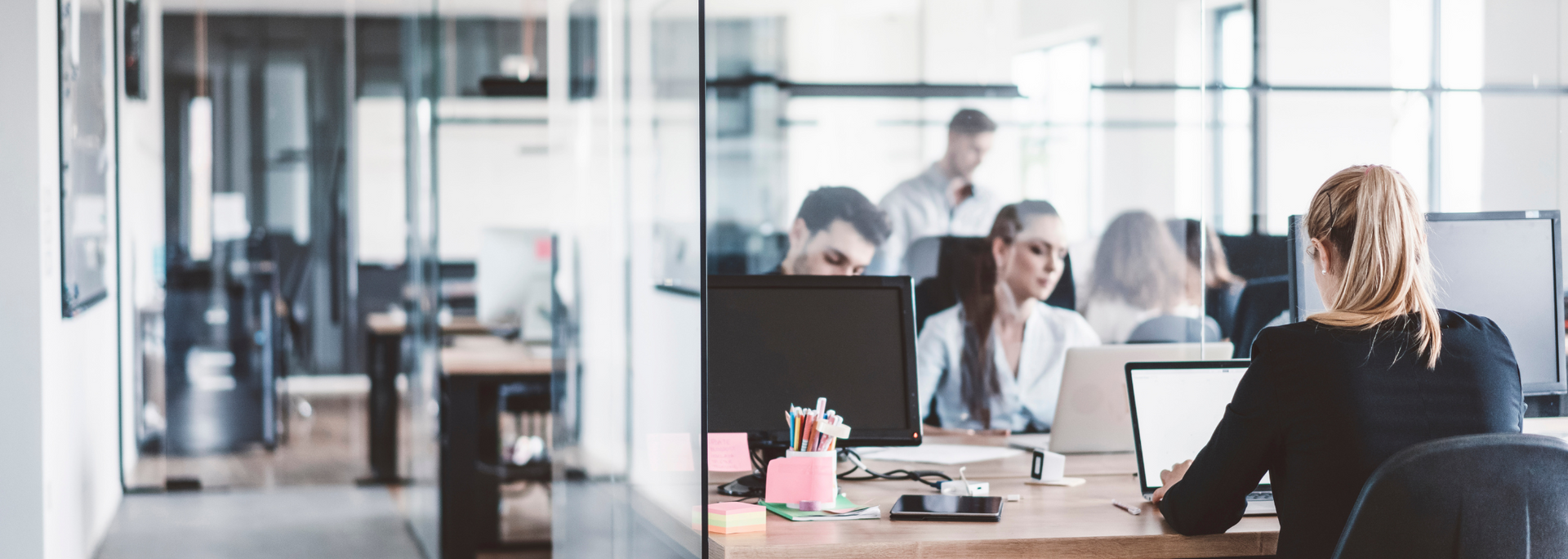 Office setting with people working at desks, some using laptops and monitors.