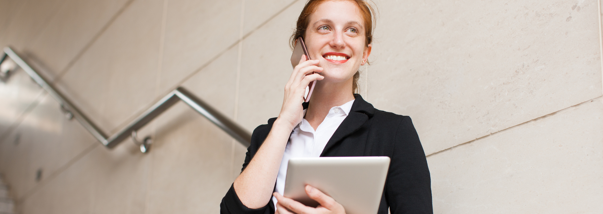 Woman on stairs smiles while on a phone call, holding a tablet. She wears a black blazer and a white shirt.