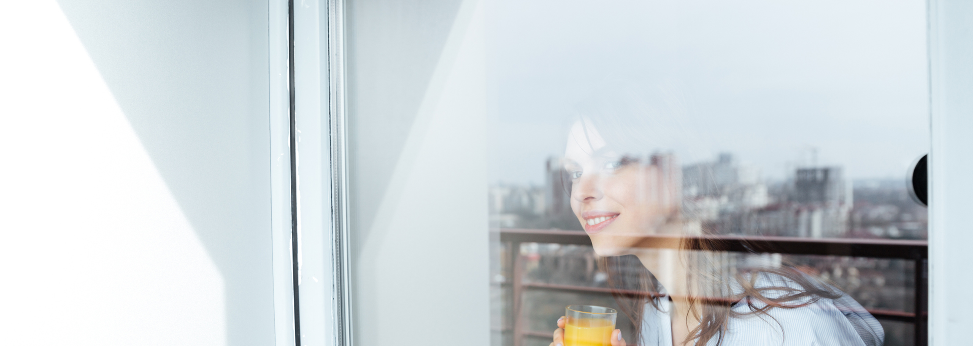 Woman smiles while looking out a window onto a cityscape with a glass of orange juice.