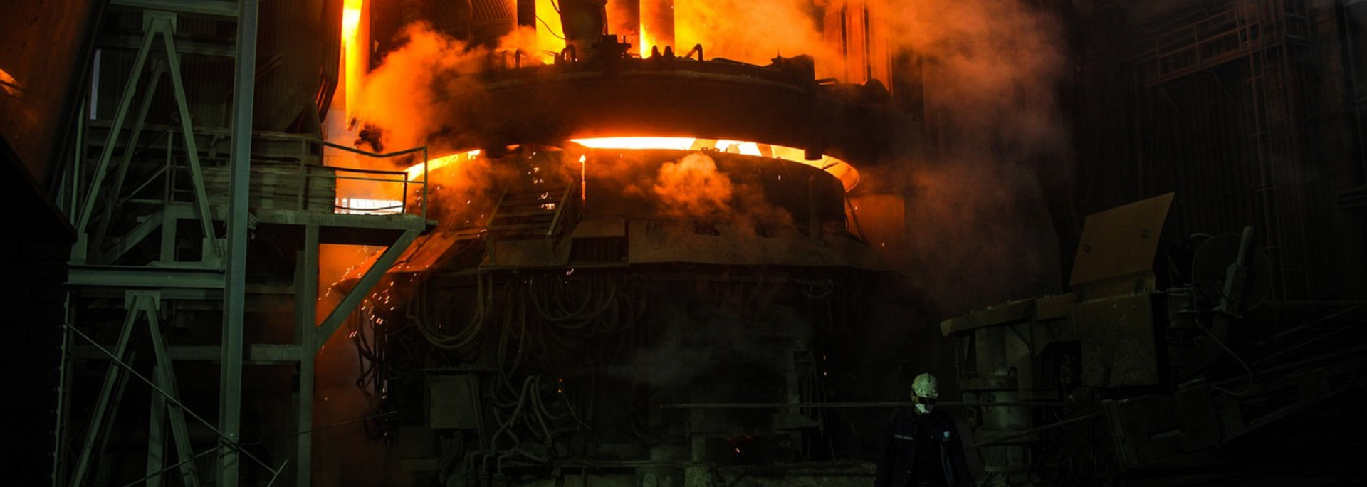 Molten metal pouring from furnace. Intense orange light, sparks, and steam fill the industrial setting.