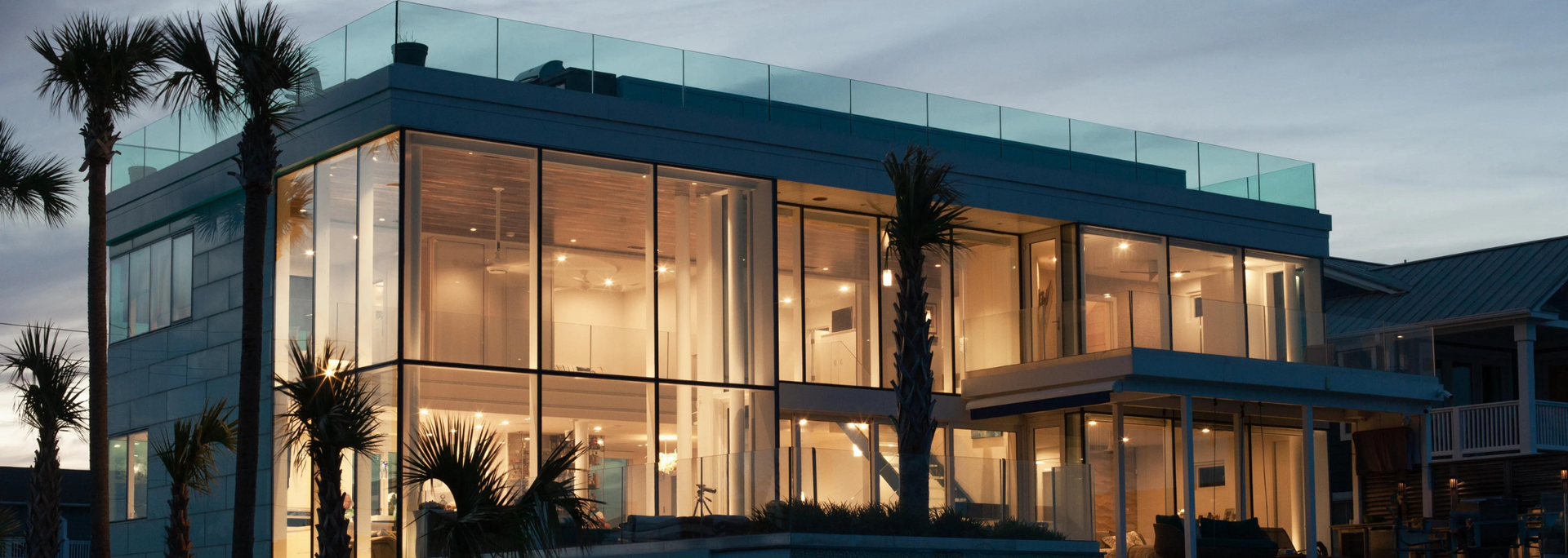 Modern building with glass walls, palm trees, and a glass-paneled roof at dusk.