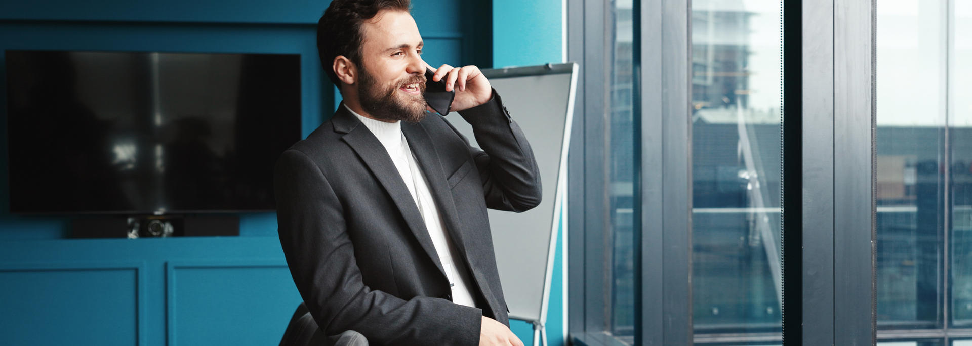 A man in a suit talks on the phone while looking out a large office window.