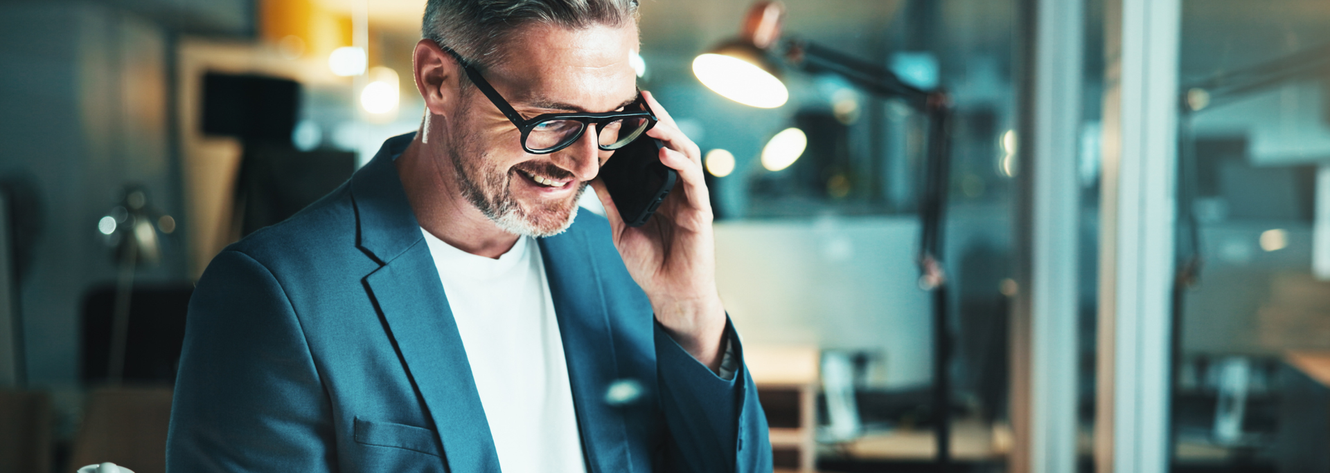 Man smiling while talking on a phone in a modern office with glass walls.