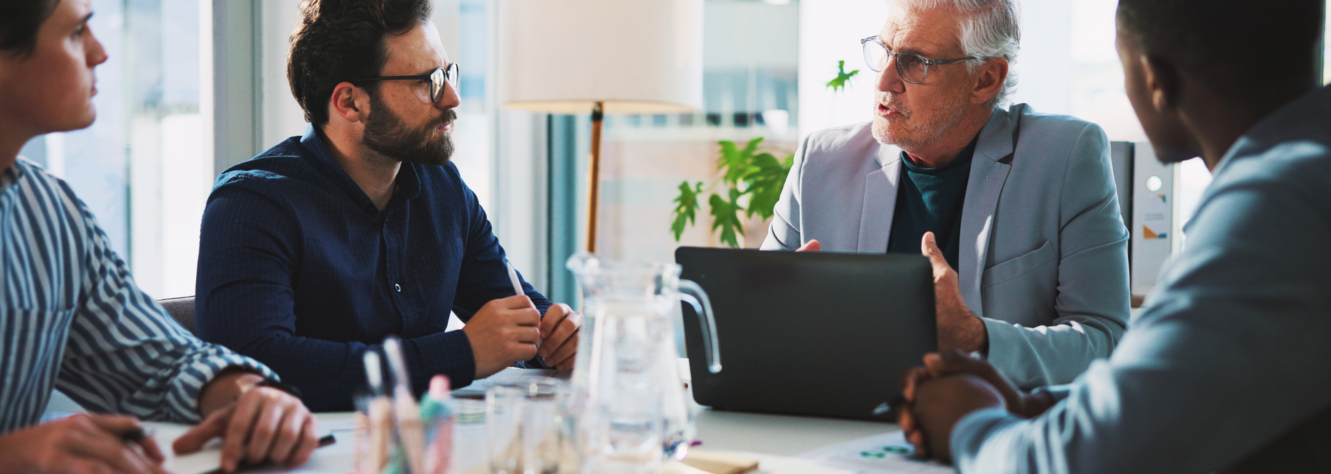 Business meeting in a bright office; four people seated around a table, one speaking and gesturing.