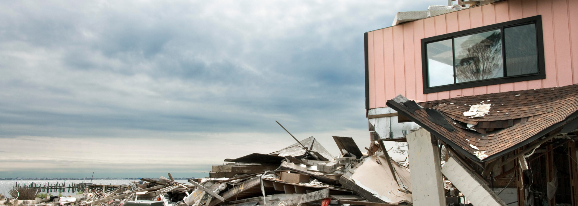 Debris of a collapsed pink building with a window, dark cloudy sky, and the ocean in the background.