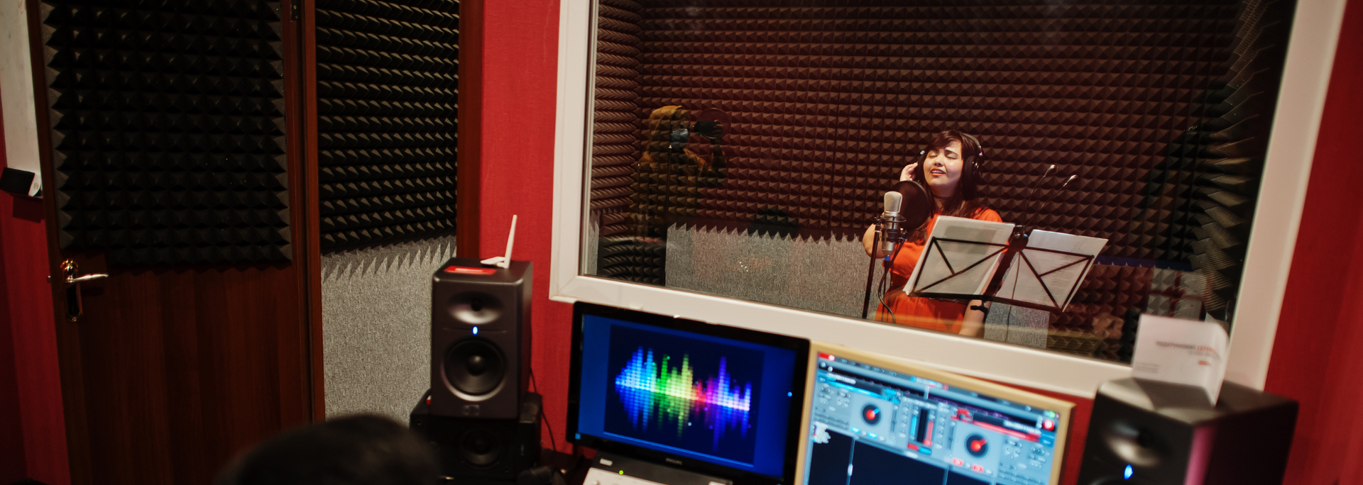 A woman records vocals in a soundproof booth. Recording equipment is visible in foreground.