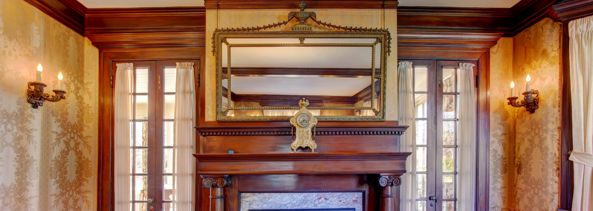 Fireplace with ornate mirror and sconces in a room with wood trim and decorative wallpaper.