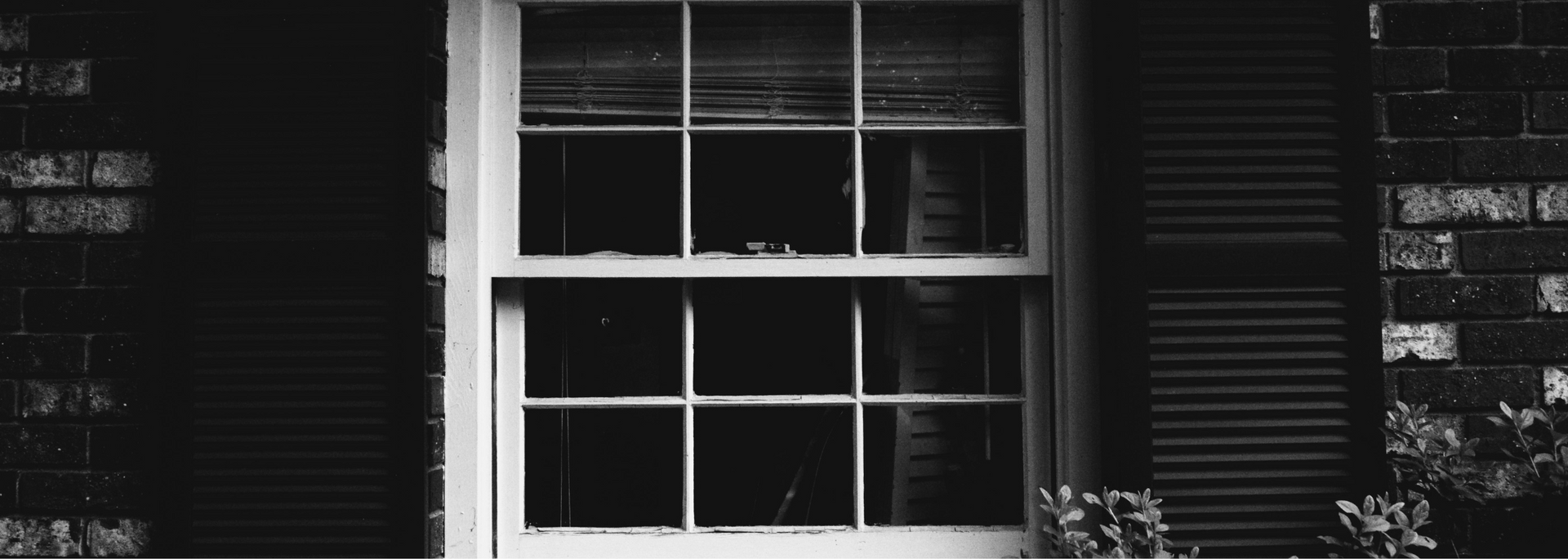 Black and white photo of a window with blinds, shutters, and brick siding.