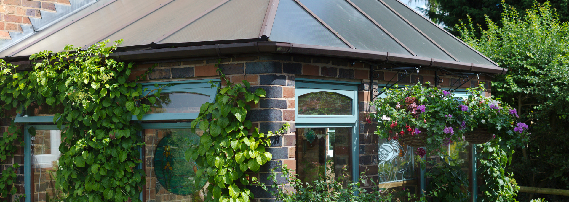 Brick greenhouse with glass roof and hanging flower baskets, covered in vines.