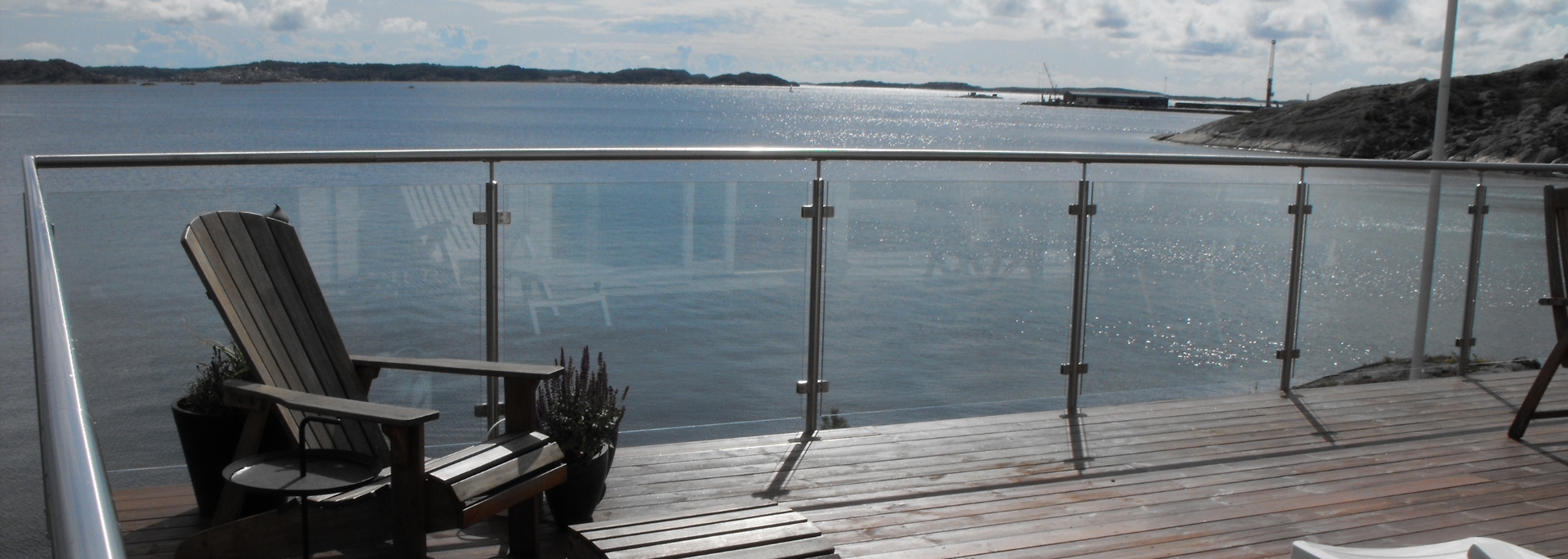 A waterside patio with a glass railing, Adirondack chair, and a view of the water and distant land.