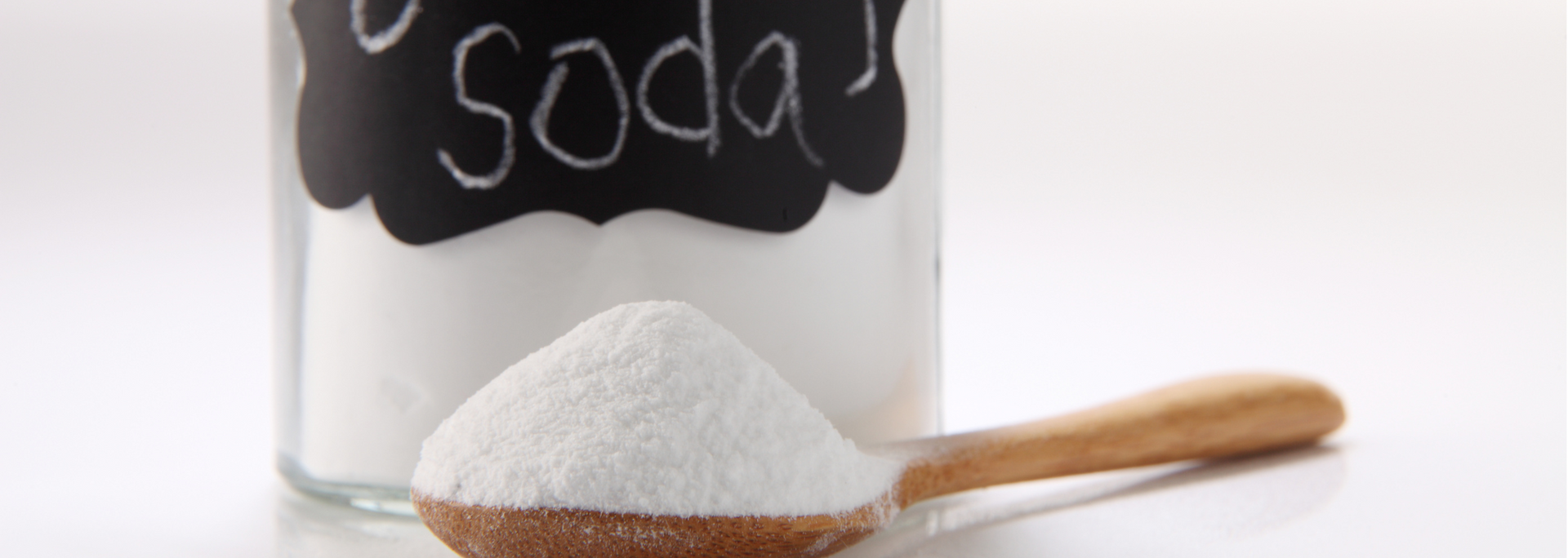 Baking soda in a jar with a wooden spoon holding a scoop of it against a white background.