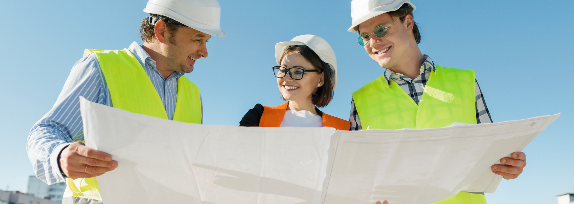 Three people in hard hats and safety vests looking at a blueprint outdoors.