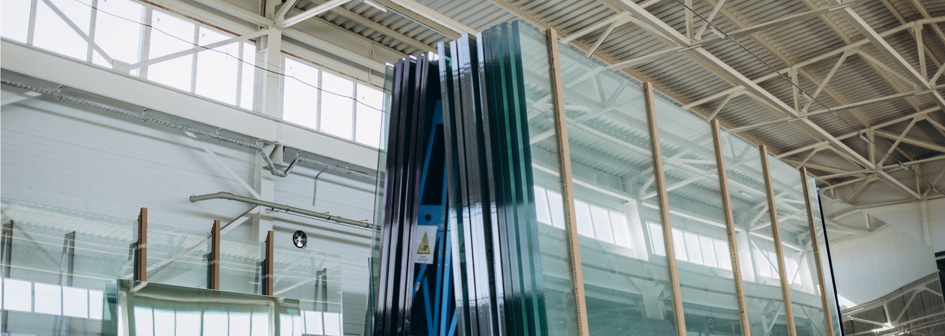 Stacks of glass panels inside a warehouse. Supported by wooden frames, under the roof's steel beams.
