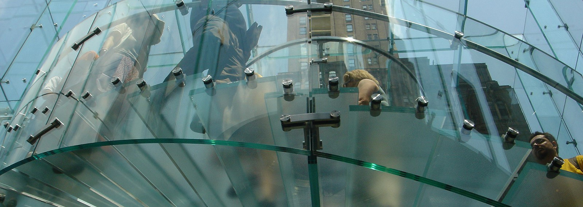 Curving glass structure with visible fasteners. Buildings reflected in the transparent surfaces.