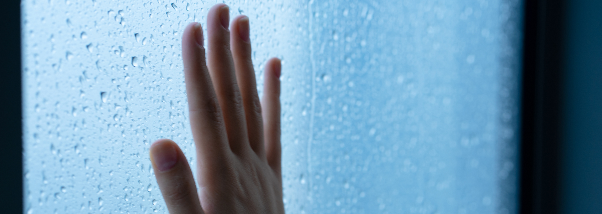Hand touching a glass surface covered in raindrops. Blue hues.