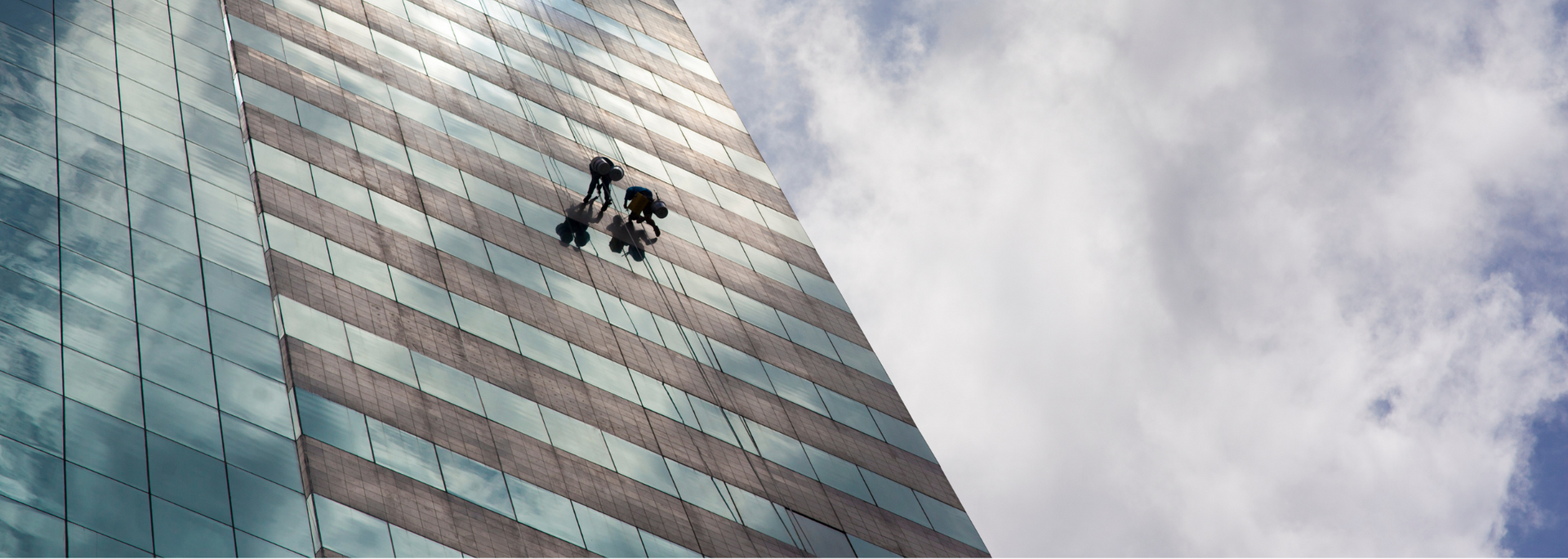 Two window washers suspended on the side of a tall building. Blue sky and white clouds visible.