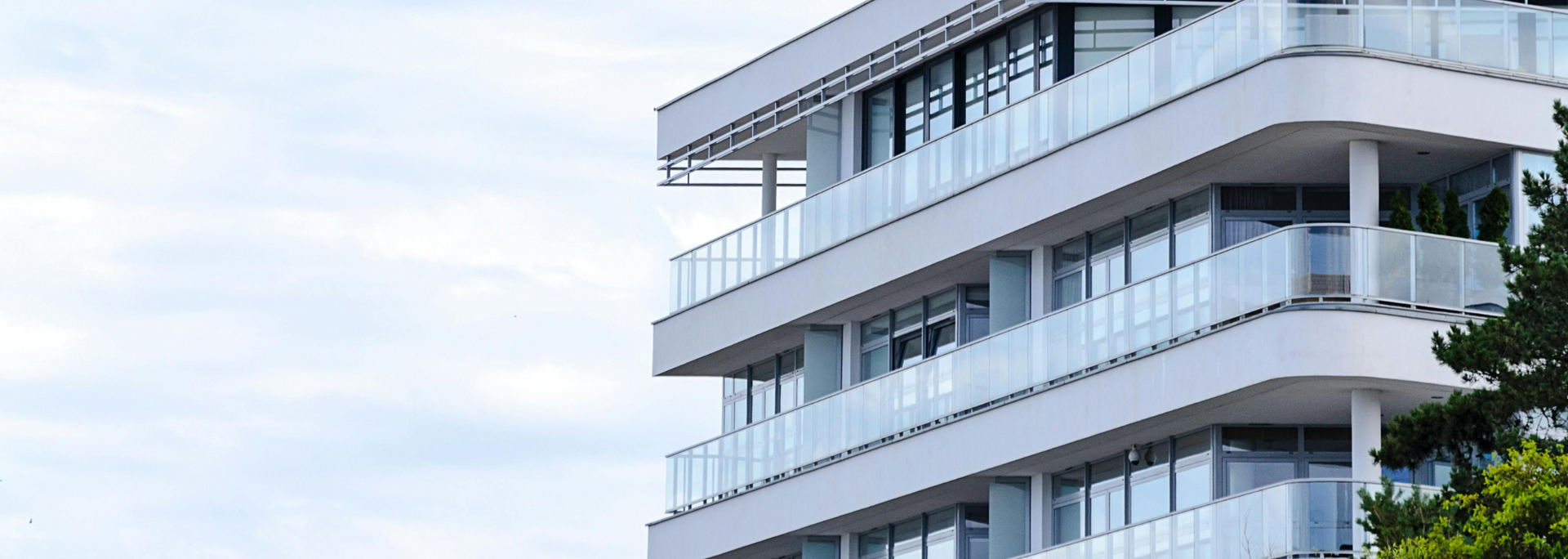 Modern white multi-story building with glass balconies against a cloudy sky.