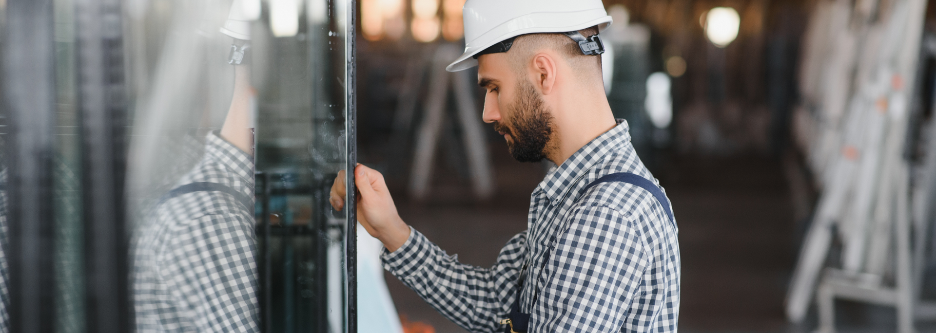 A worker in a white hard hat and plaid shirt inspects a glass panel, likely in a workshop.