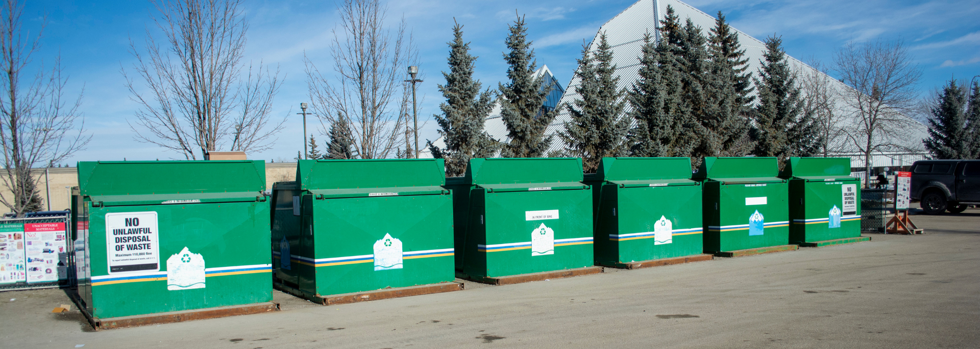Green recycling bins with white logos in front of trees and a building on a sunny day.