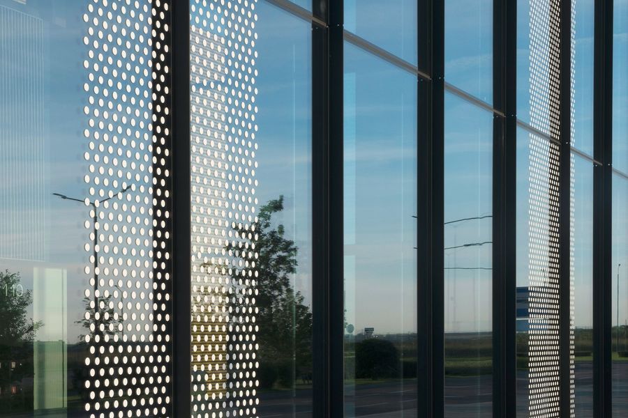 Exterior of a modern building with large glass windows and perforated panels. Reflects sky and trees.