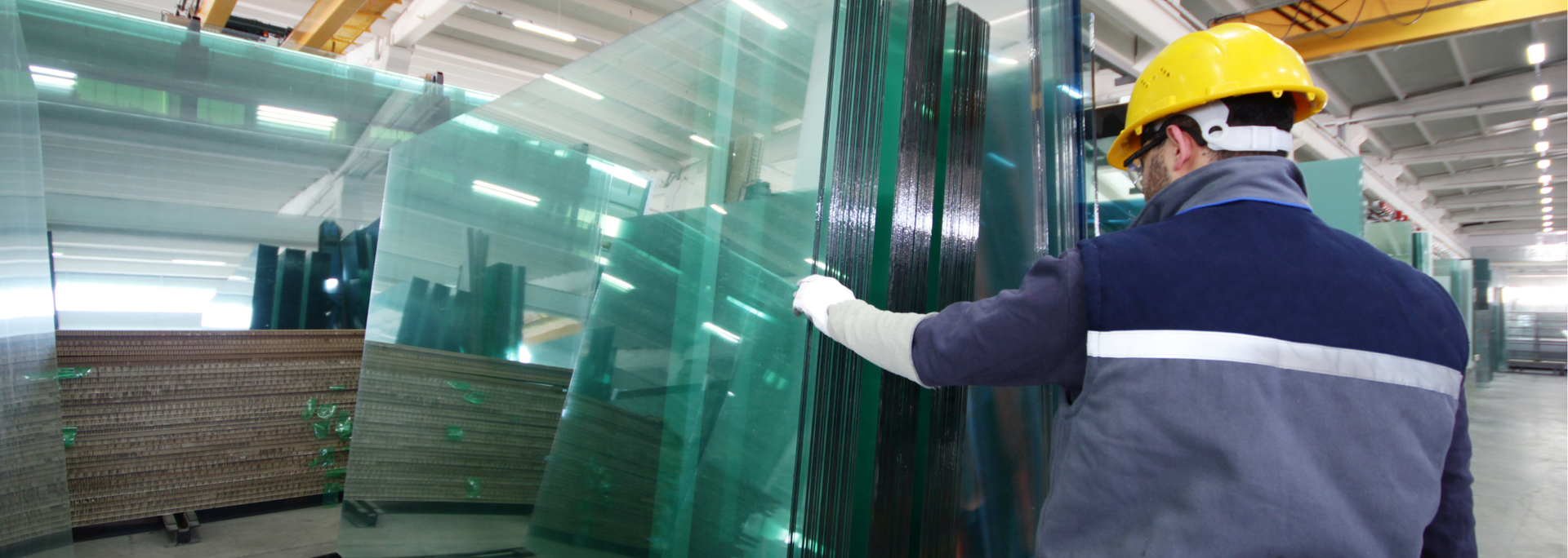 Factory worker in hard hat inspecting stacked glass sheets.