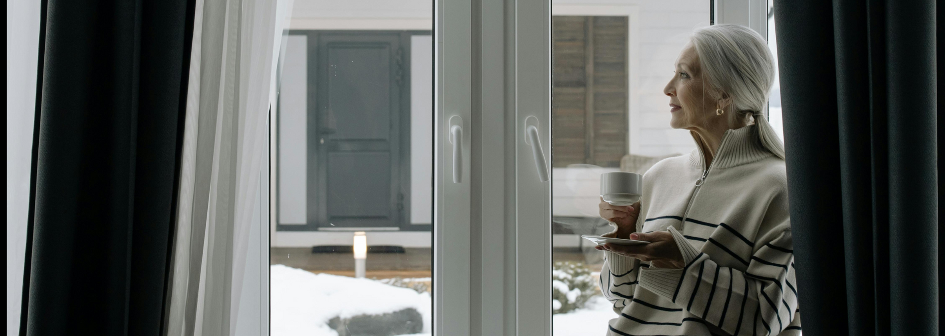 Elderly person with gray hair looking out a window while holding a mug. Black curtains frame the view.