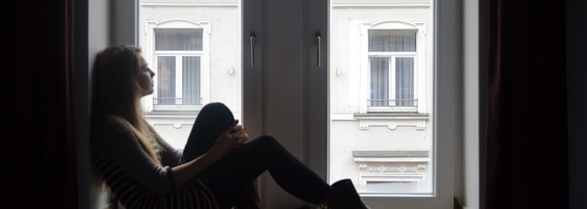 Woman sits in a window, looking outside. The setting is indoors, with natural light and a building visible outside.