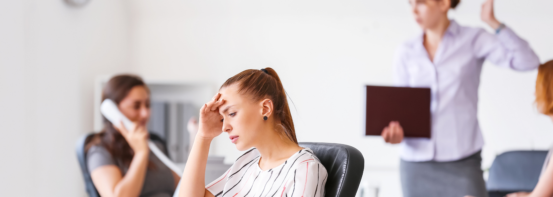 Woman with hand on forehead looks stressed in a meeting; another gestures in the background.