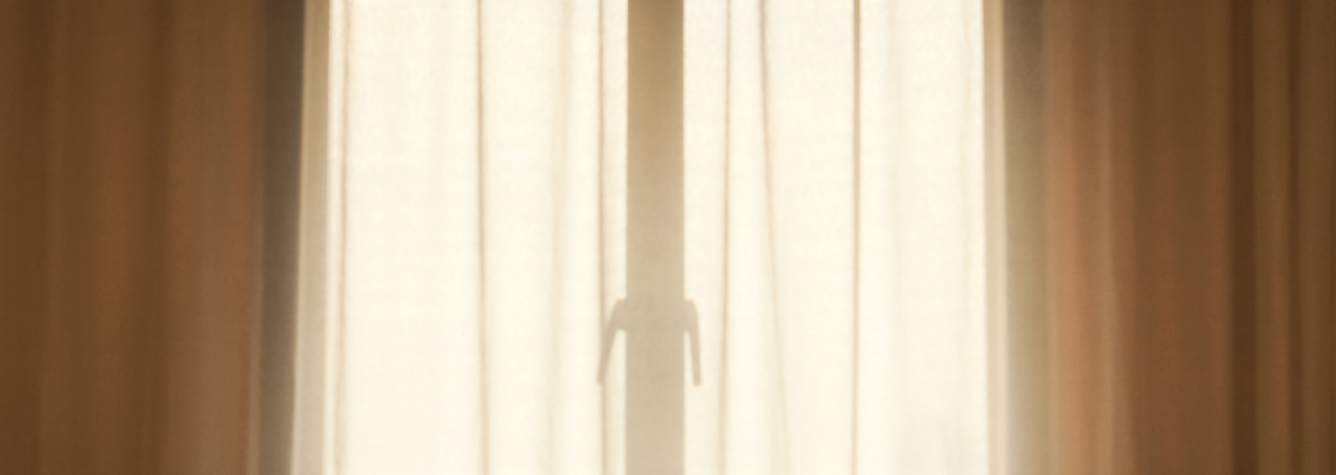 Shadow of a person standing behind sheer curtains, sunlight filtering through.